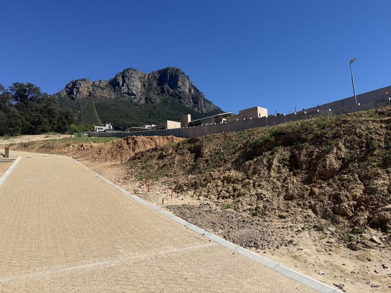 Paved road curving past a bare earth bank, with a boundary wall, buildings and a rocky mountain behind